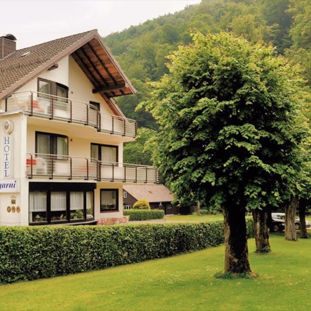 Außenansicht des Iris Valley Lodge in Herzberg am Harz, weißes Gebäude mit Balkonen vor bewaldeten Bergen, kleines Hotel mitten in der Natur des Harzes
